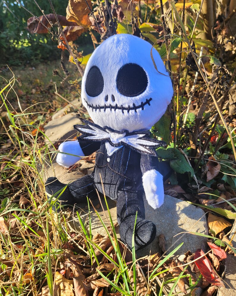 Op de afbeelding: Een pluche speelgoed van Jack Skellington uit de film Nightmare Before Christmas. Het speelgoed zit op een rots met herfstbladeren en gras eromheen. Het speelgoed draagt een zwart-wit gestreept pak met een witte kraag en een zwarte strik.
