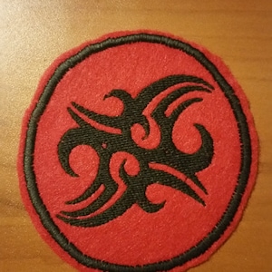 May include: A round, red patch with a black tribal design in the center. The design is surrounded by a black border. The patch is likely made of felt or a similar fabric. This patch could be used to decorate clothing or accessories.
