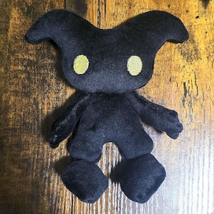 May include: A black plush doll with a unique design. It has a rounded head with two curved horns, yellow embroidered eyes, and small arms and legs. The doll is made of soft material and is sitting on a wooden surface.