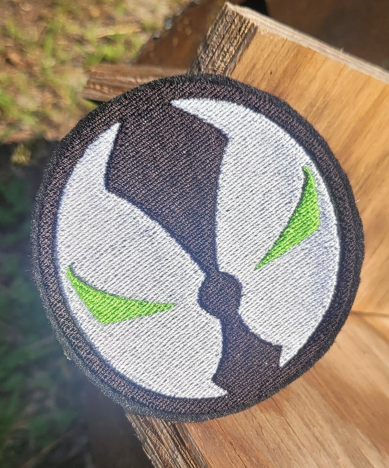May include: A black and white embroidered patch with a green symbol. The symbol is a stylized skull with two green eyes.