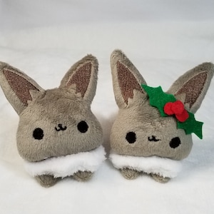 May include: Two plush toys shaped like a fox with brown ears and a white fluffy collar. One fox is wearing a green holly leaf and red berries.