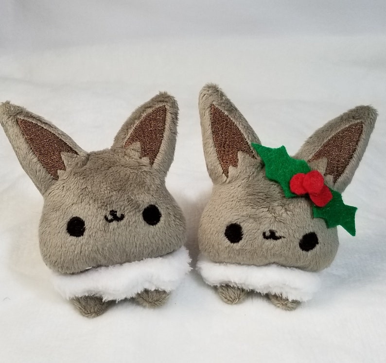 May include: Two plush toys shaped like a fox with brown ears and a white fluffy collar. One fox is wearing a green holly leaf and red berries.