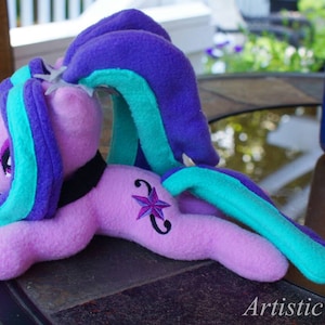 May include: A plush toy of a purple and teal pony with a pink body and a purple star on its flank. The pony is lying on its side with its head turned to the left.