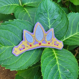 Puede incluir: Una corona bordada de color morado y dorado con un centro azul. La corona está sobre un fondo de hojas verdes.