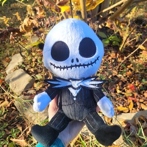 Op de afbeelding: Een pluche speelgoed van Jack Skellington uit de film Nightmare Before Christmas. Het speelgoed draagt een zwart-wit gestreept pak met een witte kraag en een zwarte strik. Het speelgoed heeft een wit gezicht met zwarte ogen en een gestikte glimlach.