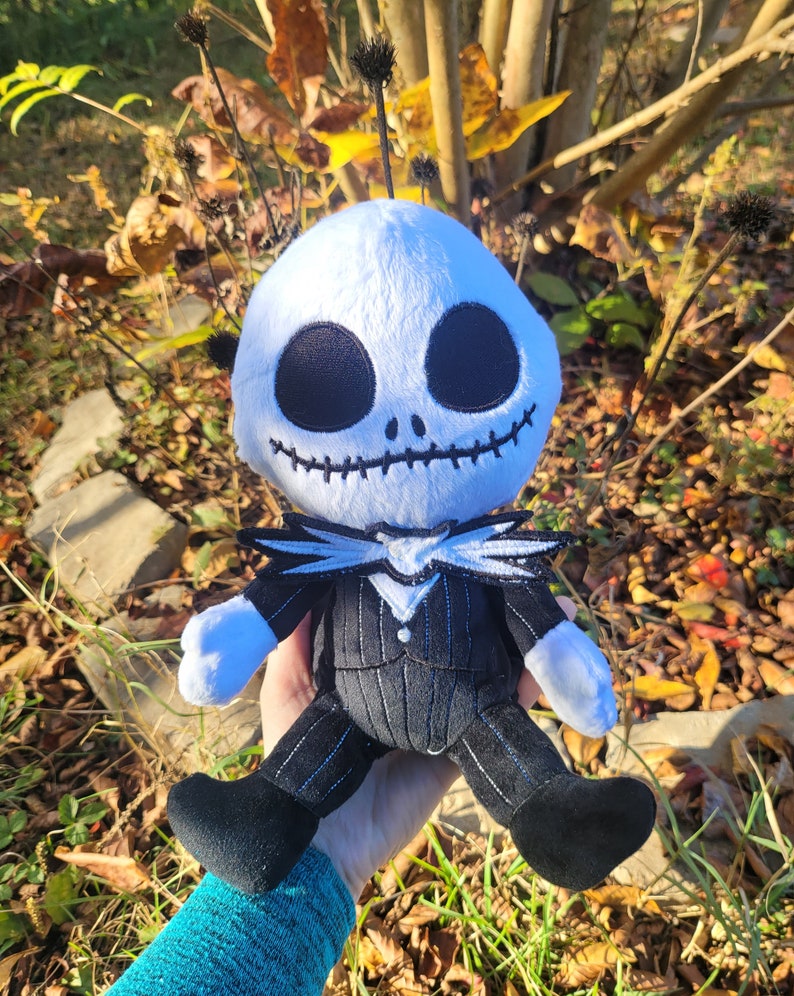 Op de afbeelding: Een pluche speelgoed van Jack Skellington uit de film Nightmare Before Christmas. Het speelgoed draagt een zwart-wit gestreept pak met een witte kraag en een zwarte strik. Het speelgoed heeft een wit gezicht met zwarte ogen en een gestikte glimlach.