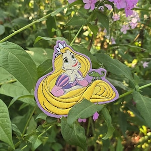 Puede incluir: Un parche bordado con Rapunzel de la película de Disney Enredados. Tiene el pelo largo y rubio, un vestido morado y una corona. Está sentada con su camaleón mascota, Pascal.