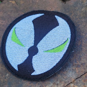 May include: A black and white embroidered patch with a green symbol. The patch is circular and has a white background with a black outline. The symbol is a stylized face with green eyes.