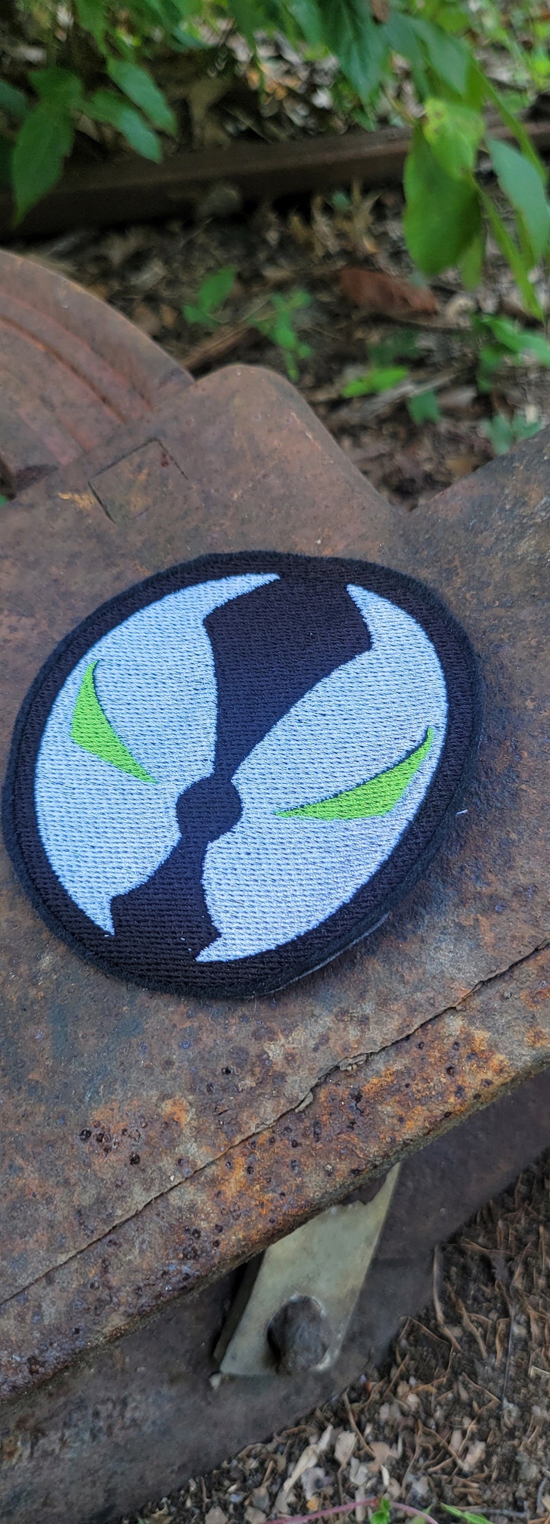 May include: A black and white embroidered patch with a green symbol. The patch is circular and has a white background with a black outline. The symbol is a stylized face with green eyes.