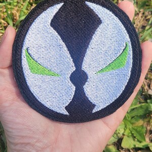May include: A black and white embroidered patch with a green symbol. The symbol is a stylized face with two green eyes. The patch is circular and has a black border.