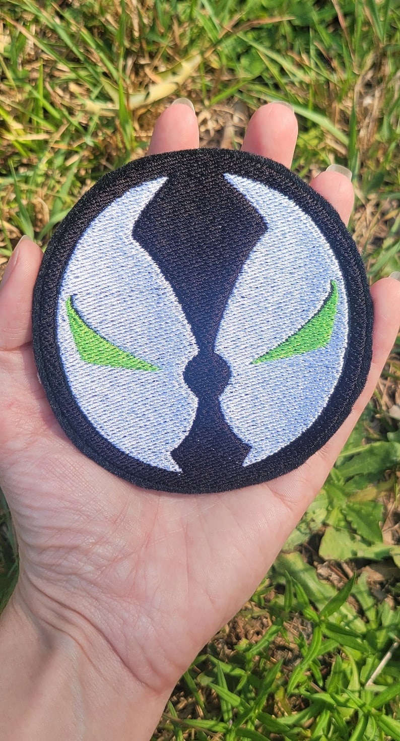 May include: A black and white embroidered patch with a green symbol. The symbol is a stylized face with two green eyes. The patch is circular and has a black border.