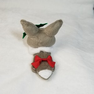 May include: A gray plush toy shaped like a fox wearing a white collar and a red bow. The fox has green holly leaves on its head.