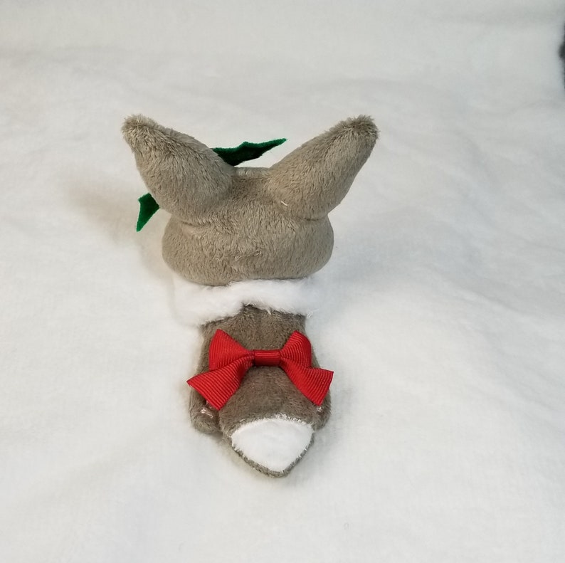 May include: A gray plush toy shaped like a fox wearing a white collar and a red bow. The fox has green holly leaves on its head.