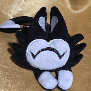 May include: A plush black cat toy with white accents on the face, paws, and ears. The cat has a white smiling face and a long tail with a white tip. The toy is lying on a gold-colored surface.