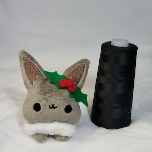 May include: A small gray plush toy with a white fur collar and a green holly leaf with red berries on its head.