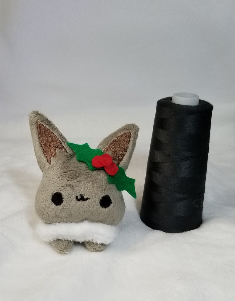 May include: A small gray plush toy with a white fur collar and a green holly leaf with red berries on its head.