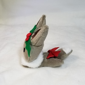 May include: A gray plush bunny with a white fur collar, wearing a green holly leaf and red berry crown and a red bow on its tail.