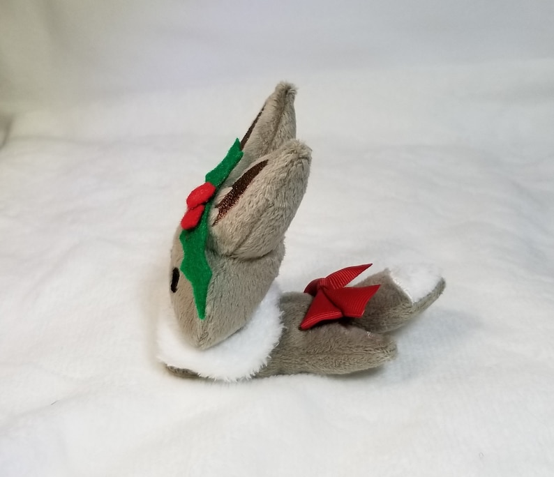 May include: A gray plush bunny with a white fur collar, wearing a green holly leaf and red berry crown and a red bow on its tail.