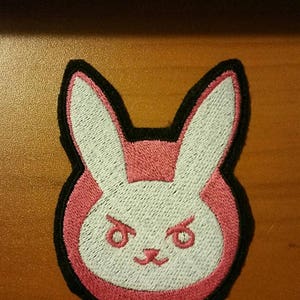 May include: A pink and white embroidered patch of a bunny rabbit with black outlining. The bunny has a grumpy expression with small, black eyes and a white nose.