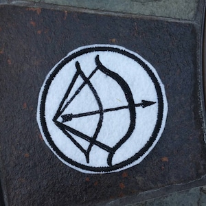 May include: White fabric patch with a black embroidered design of a bow and arrow.