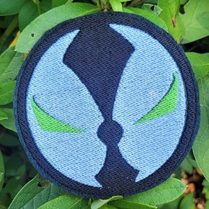 May include: A blue and black embroidered patch with a green symbol. The symbol is a stylized face with two green eyes.