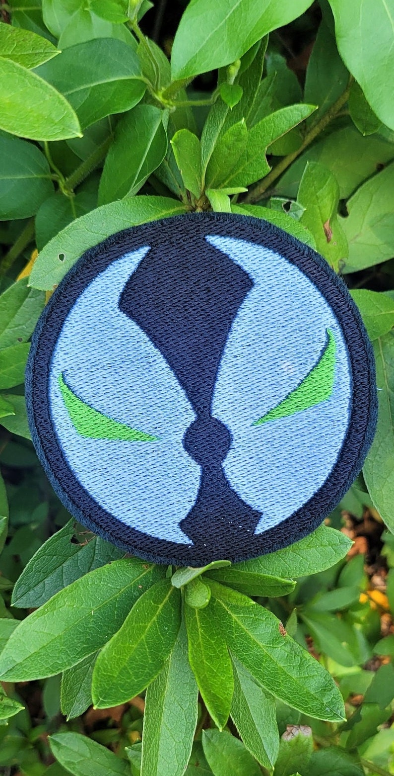 May include: A blue and black embroidered patch with a green symbol. The symbol is a stylized face with two green eyes.