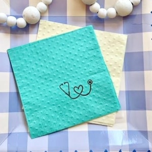 May include: A blue and white checkered paper plate with a scalloped edge. Two napkins are on the plate, one is yellow and the other is turquoise with a black stethoscope and heart design. The text "Selfish Necessities" is printed on the bottom of the plate.
