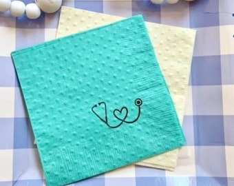 Puede incluir: Un plato de papel a cuadros azul y blanco con un borde festoneado. Dos servilletas están en el plato, una es amarilla y la otra turquesa con un estetoscopio negro y un diseño de corazón. El texto "Selfish Necessities" está impreso en la parte inferior del plato.