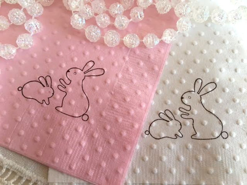 May include: Pink and white dotted napkins with black outline drawings of two bunnies. A string of clear, round beads is draped across the napkins. The napkins are likely for Easter or spring celebrations.