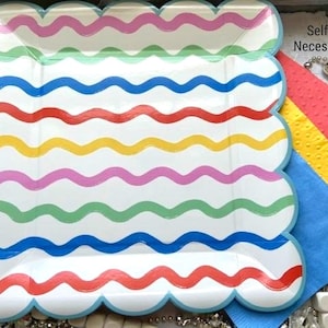 May include: A square paper plate with scalloped edges featuring horizontal wavy lines in pink, blue, red, yellow, and green. The text Selfish Necessities is printed in the upper right corner. Beside the plate are red, yellow, and blue napkins.