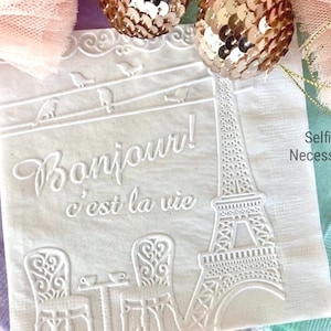 May include: White paper napkins with an embossed design of the Eiffel Tower, birds, and the text "Bonjour! c'est la vie". The napkins are decorated with a white lace border and the text "Selfish Necessities".