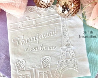 May include: A white paper napkin featuring an embossed design of the Eiffel Tower, two chairs, a small table, and birds on a wire. The text Bonjour! c'est la vie is embossed on the surface. The text Selfish Necessities is printed in black.
