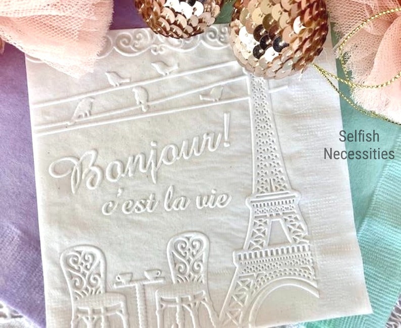 May include: White paper napkins with an embossed design of the Eiffel Tower, birds, and the text "Bonjour! c'est la vie". The napkins are decorated with a white lace border and the text "Selfish Necessities".