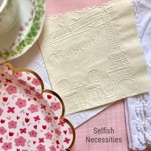 May include: A white paper napkin with an embossed design of the Eiffel Tower and the text "Paris c'est la vie". The napkin is on a pink surface with a white lace doily and a silver fork.