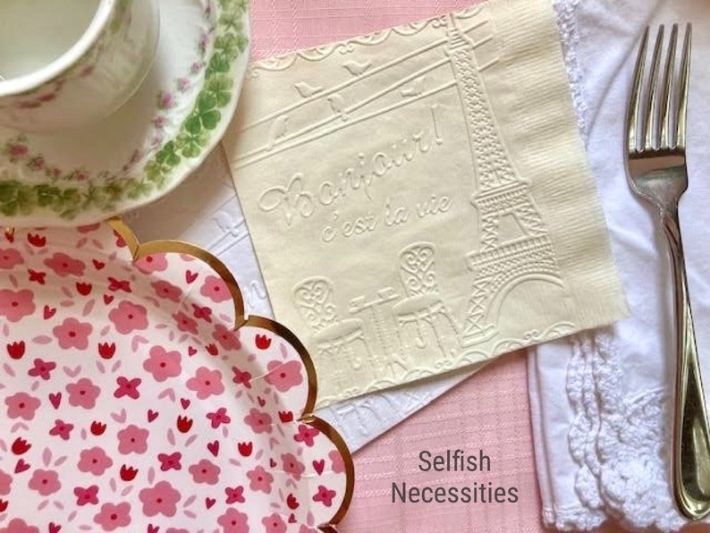 May include: A white paper napkin with an embossed design of the Eiffel Tower and the text "Paris c'est la vie". The napkin is on a pink surface with a white lace doily and a silver fork.