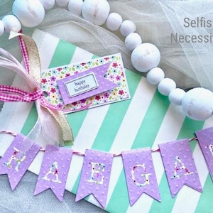 May include: A festive birthday gift presentation. The scene includes a mint green and white striped gift bag, a floral-patterned tag with "happy birthday" text, and a banner spelling out a name. A white bead garland and pink ribbon complete the arrangement.