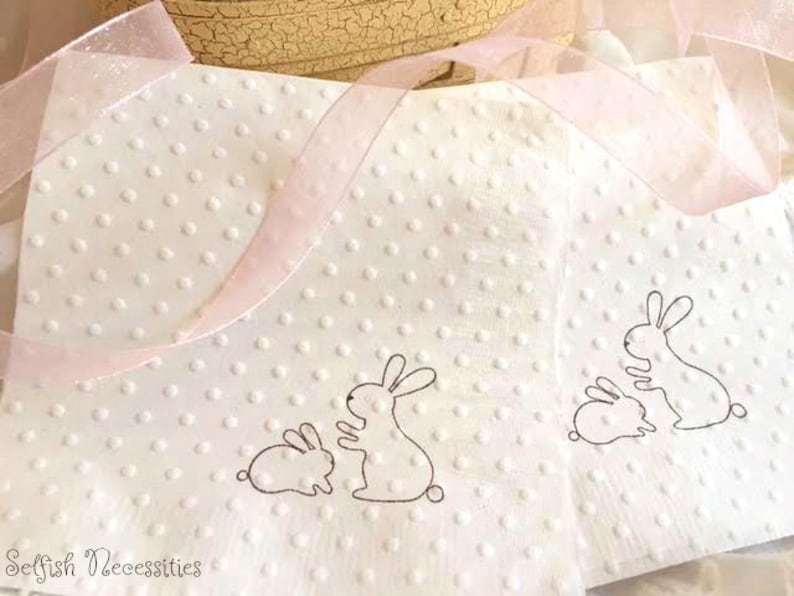 May include: White napkins with a dotted texture and bunny illustrations. The napkins have a pink ribbon and are suitable for Easter or spring celebrations. The text "Selfish Necessities" is visible in the bottom left corner.