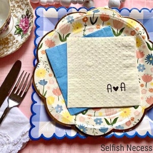 Peut inclure: Une serviette en papier blanche avec un motif floral sur une assiette à carreaux bleus et blancs. La serviette porte les lettres "A ♥ A" écrites en noir.
