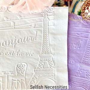 May include: Two white paper napkins with an embossed design of the Eiffel Tower and the text "Bonjour! c'est la vie". The napkins are next to each other and the one on the right is a light purple color.