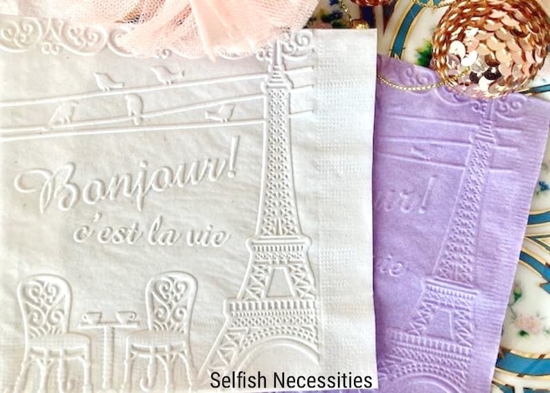 May include: Two white paper napkins with an embossed design of the Eiffel Tower and the text "Bonjour! c'est la vie". The napkins are next to each other and the one on the right is a light purple color.