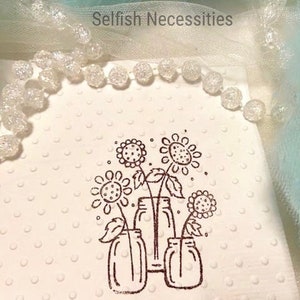 May include: A white, textured card with a brown ink illustration of sunflowers in mason jars. The card is adorned with a string of clear, spherical beads. The text "Selfish Necessities" is visible in the upper right corner.