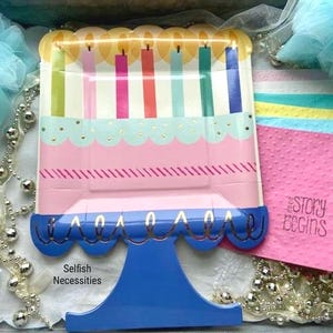 May include: A blue and white cake-shaped paper plate with candles and gold trim. The plate is on a blue stand with the words "Selfish Necessities" printed below. The plate is surrounded by white lace, silver beads, and pink and blue napkins with the words "The Story Begins" printed on one.