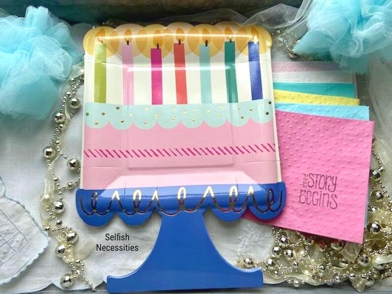 May include: A blue and white cake-shaped paper plate with candles and gold trim. The plate is on a blue stand with the words "Selfish Necessities" printed below. The plate is surrounded by white lace, silver beads, and pink and blue napkins with the words "The Story Begins" printed on one.