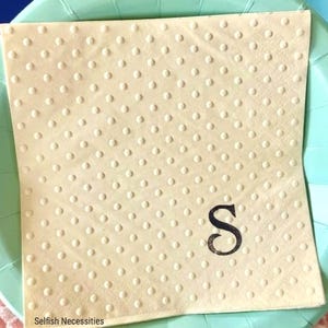 May include: A square, cream-colored paper napkin featuring an embossed polka dot pattern and a single, black, serif-style letter S printed in the lower right corner. The text Selfish Necessities is printed in small black font at the bottom left.
