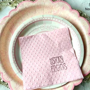 May include: A pink paper napkin with white polka dots and the text "and the story begins" in brown lettering. The napkin is on top of two pink and white paper plates with scalloped edges.