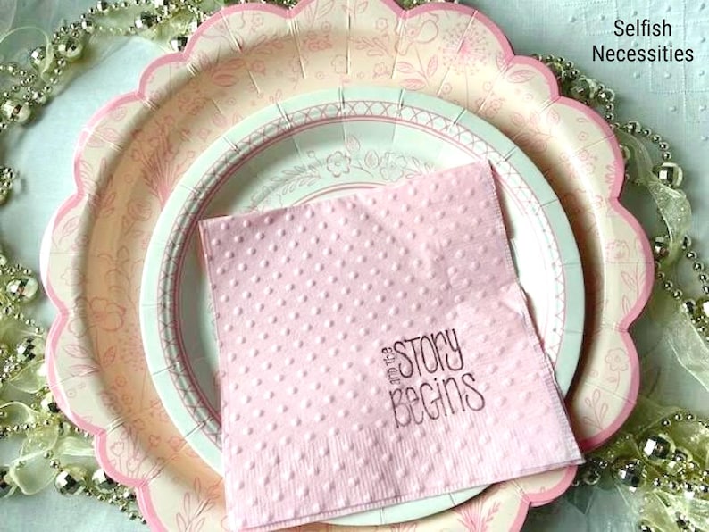 May include: A pink paper napkin with white polka dots and the text "and the story begins" in brown lettering. The napkin is on top of two pink and white paper plates with scalloped edges.