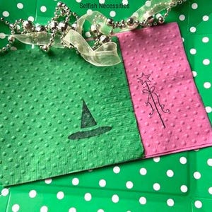 May include: Two green and pink paper napkins with a witch's hat and a magic wand design. The napkins are on a green and white polka dot surface. The text "Selfish Necessities" is visible in the top left corner of the image.