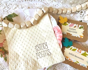 May include: A cream-colored textured napkin with the black printed text 'and the STORY BEGINS', accompanied by two brown rectangular place cards featuring leaf cutouts and the text 'Custom Place Cards' and 'Selfish Necessities', with wooden beads.