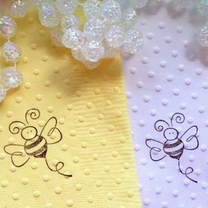 Bumble bee image hand stamped on pale yellow and white napkins. The been has a swirl below indicating that it is flying. The bees wings are out stretched and also has curly antennae. Napkins are textured with polka dot embossing.