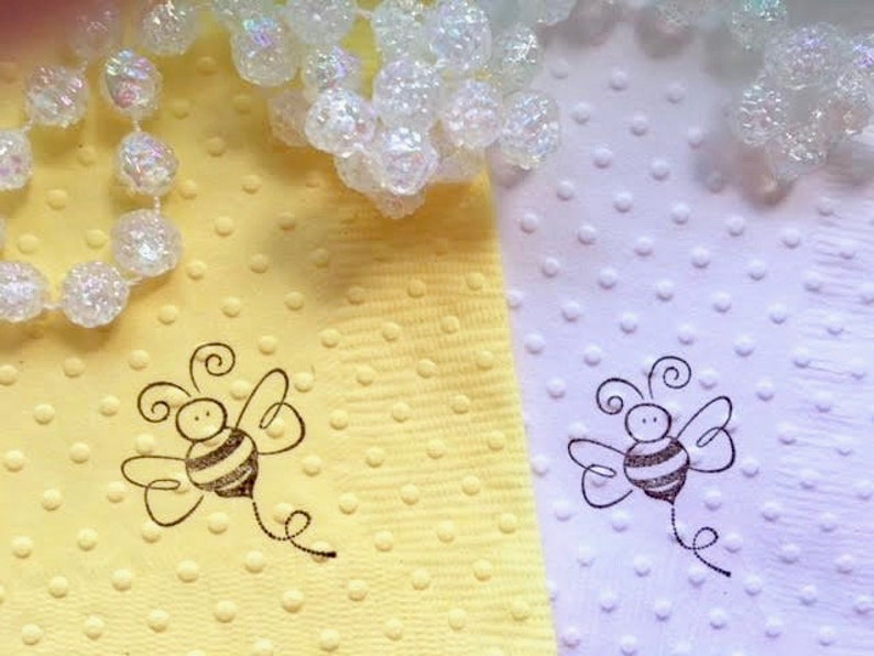 Bumble bee image hand stamped on pale yellow and white napkins. The been has a swirl below indicating that it is flying. The bees wings are out stretched and also has curly antennae. Napkins are textured with polka dot embossing.